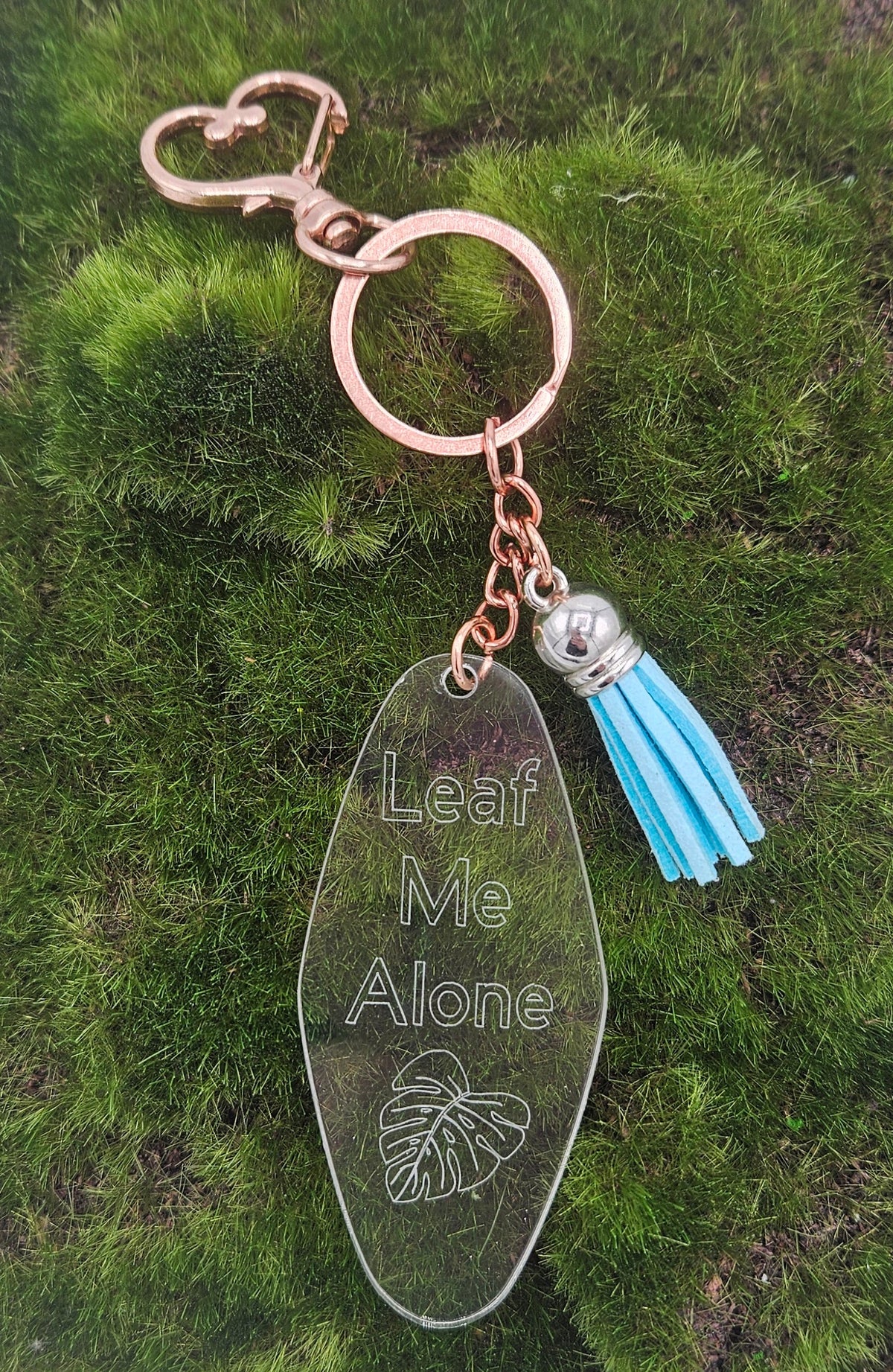 A rose gold keychain with a clear acrylic pendant engraved with 'Leaf Me Alone' and a light blue tassel.