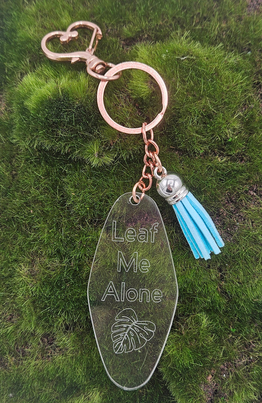 A rose gold keychain with a clear acrylic pendant engraved with 'Leaf Me Alone' and a light blue tassel.
